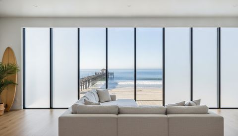 Huntington Beach living room with a wall of switchable smart glass panels facing the pier — alternating panels clear over the sand and the Pacific, the rest switched to flat, evenly frosted milky-white privacy