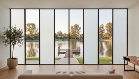 Irvine great room in Woodbridge with a wall of eight switchable smart glass panels facing the lake and dock — alternating panels clear to the water and sycamores, the rest switched to flat, evenly frosted milky-white privacy