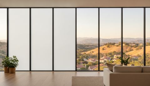 Santa Clarita hillside great room at late afternoon with a west-facing wall of floor-to-ceiling switchable smart glass — the left panels switched to flat, even milky-white privacy, the right panels clear to golden chaparral hills, tile rooftops and the mountains beyond